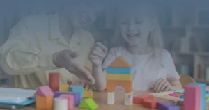 Criança sorridente realizando atividade com blocos de montar durante avaliação psicológica, acompanhada por profissional em ambiente terapêutico.