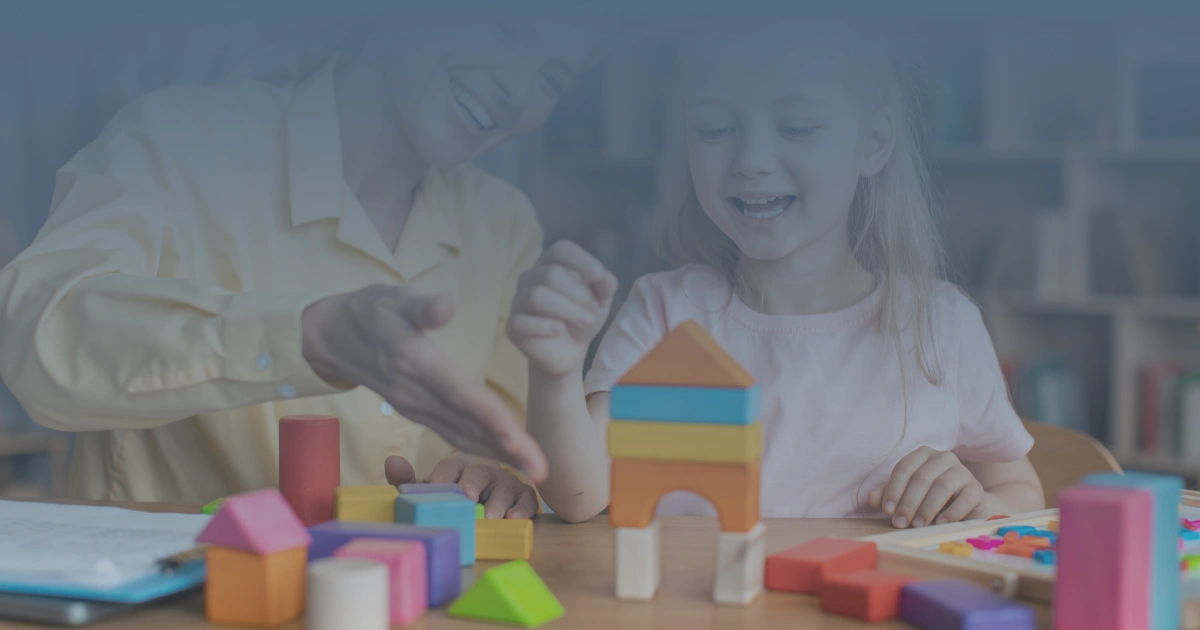 Criança sorridente realizando atividade com blocos de montar durante avaliação psicológica, acompanhada por profissional em ambiente terapêutico.